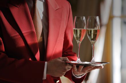 Ein Kellner in roter Kellneruniform trägt zwei gefüllte Champagnergläser auf einem Tablett im Hotel Baiersbronn