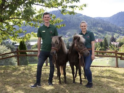 Zwei Ponys mit Pflegern stehen vor einer bezaubernden Waldlandschaft im Schwarzwald